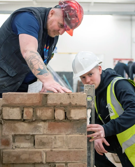 A brickwork lecturer guiding a student as they carefully check alignment and leveling of a brick wall construction in a practical workshop. A brickwork lecturer guiding a student as they carefully check alignment and leveling of a brick wall construction in a practical workshop.